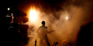 Guitarist performing on stage with crowd and bright lights.