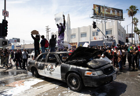 Protesters standing on damaged police car, holding signs.