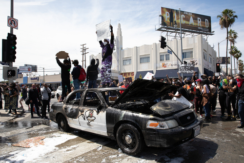 Protesters standing on damaged police car, holding signs.