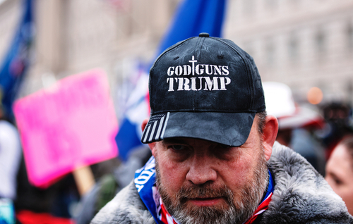 shutterstock_1888591831 (1).jpg featured image Man wearing God Guns Trump hat in a crowd.