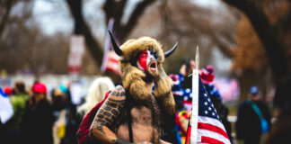 Man wearing horned fur hat, painted face, holding American flag.