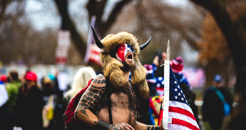 Man wearing horned fur hat, painted face, holding American flag.