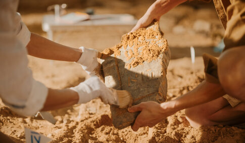 Two individuals carefully excavating a large stone artifact at an archaeological site