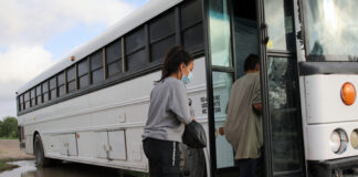 People boarding a white bus on a dirt road.