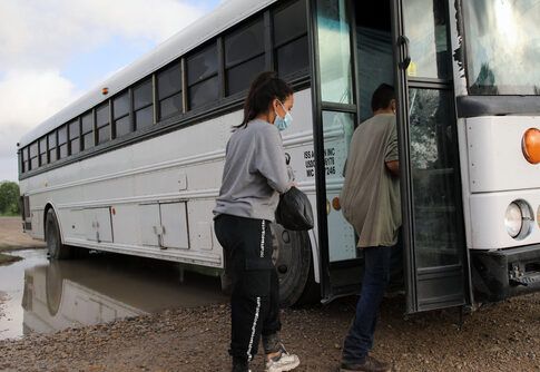 People boarding a white bus on a dirt road.
