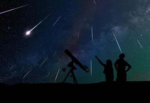 Silhouetted people stargazing with telescope under meteor shower.