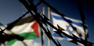 Barbed wire with Palestinian and Israeli flags blurred in background.