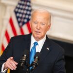 Man speaking at podium with American flag in background.