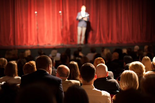 shutterstock_495240280.jpg featured image Person on stage speaking to seated audience in theater.