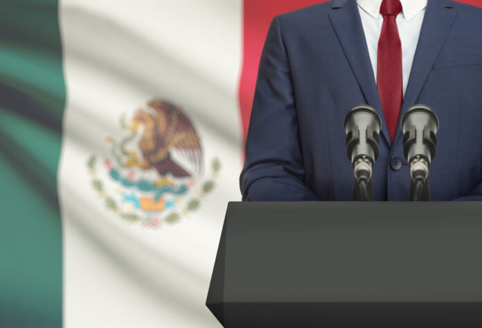 Man in suit with Mexican flag speaking at podium